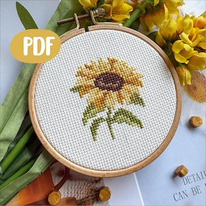 May include: A cross-stitch sunflower design in a wooden embroidery hoop. The sunflower features yellow and brown hues, with green leaves and stem. The word "PDF" is in a yellow oval. The craft project is displayed with flowers and decorative elements.