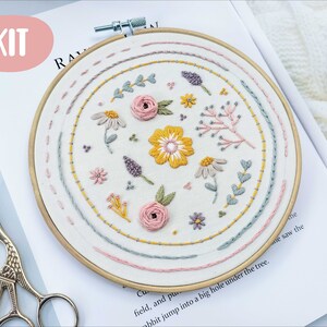 Könnte beinhalten: Stickset mit floralem Design in einem Holzreifen. Das Design umfasst rosa, gelbe und lavendelfarbene Blumen und grüne Blätter, die auf weißen Stoff gestickt sind. Das Wort "KIT" befindet sich in einem rosa Kreis.