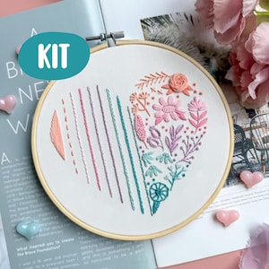 Op de afbeelding: Borduurpakket met een hartontwerp met kleurrijke bloemen- en lineaire patronen. Het hart is geborduurd met roze, perzik, teal en paarse draden. De set bevat een houten borduurring en het woord "KIT" in een teal ovaal.