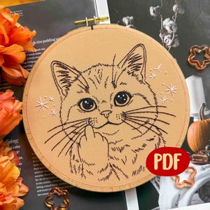 Peut inclure: Broderie d'un visage de chat dans un cercle en bois. Le chat fait un doigt d'honneur. La broderie est en fil noir sur un tissu beige. Des éclats d'étoiles blancs sont cousus autour de la tête du chat. Un logo PDF rouge est en bas à droite.