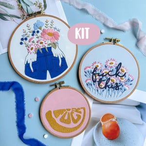 以下が含まれることがあります： 刺繍枠のアート作品が3つ。1つは手と青いジーンズのフローラルデザイン、もう1つは花と一緒に「fuck this」と書かれており、最後はピンクの背景に黄色の柑橘系のスライスがあります。「KIT」という文字も見えます。
