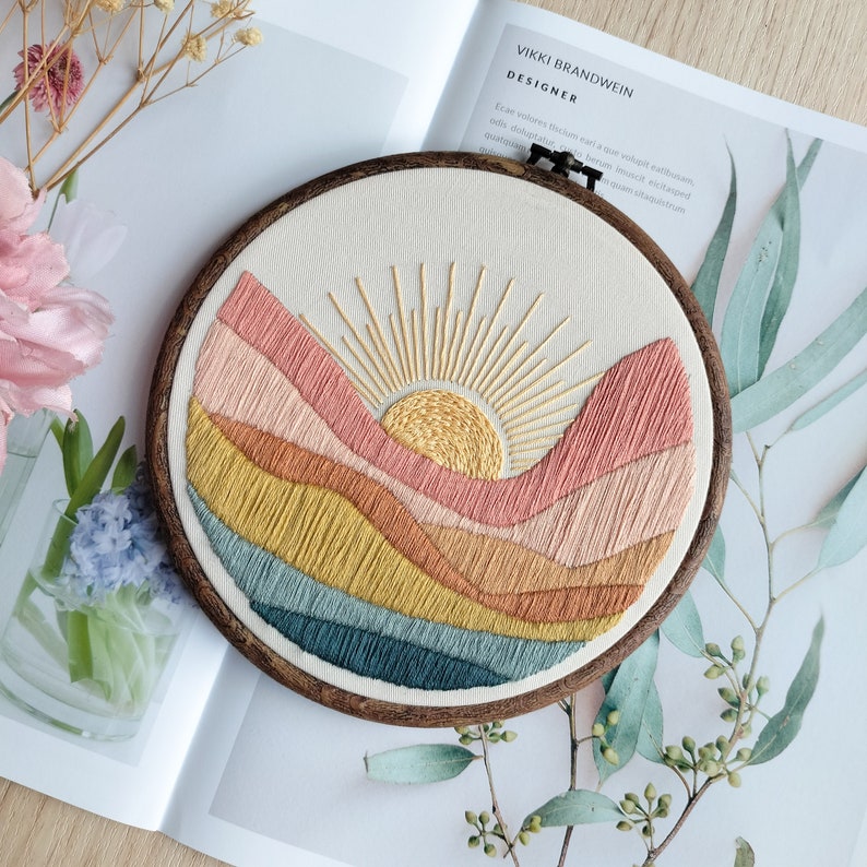 May include: Embroidered hoop art featuring a sunset over a landscape of colorful hills. The sun is yellow with gold rays and the hills are shades of pink, orange, yellow, and blue.