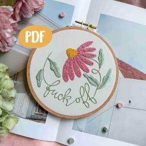 Könnte beinhalten: Ein bestickter Blumenkranz mit einer rosa und gelben Gänseblümchen-Stickerei, grünen Blättern und dem Schriftzug "fuck off" in grüner Schrift. Das Design ist in einem Holzrahmen gefasst, mit einem PDF-Label.