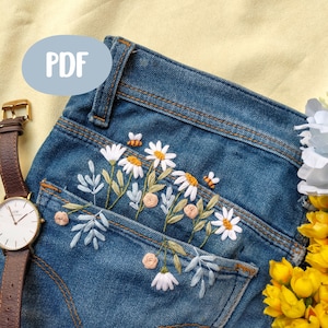 Puede incluir: Un par de pantalones vaqueros azules con un bolsillo bordado con margaritas blancas, hojas verdes y pequeñas flores rosas y amarillas.