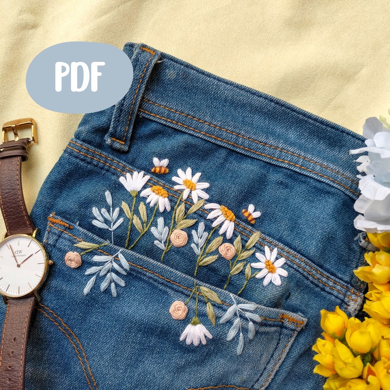 Peut inclure: Un jean bleu denim avec une poche brod&eacute;e de marguerites blanches, de feuilles vertes et de petites fleurs roses et jaunes.
