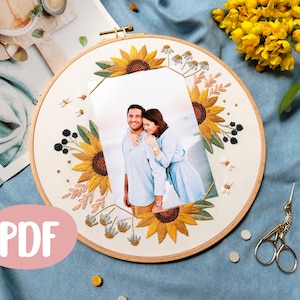 以下が含まれることがあります： 中央にカップルが写った写真が入った刺繍のフープアート。フープはひまわり、葉、ベリーで飾られています。背景は薄いブルーの布地です。