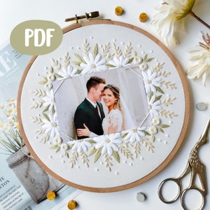 May include: A white embroidery hoop with a photo of a couple inside a wreath of white flowers and green leaves. The hoop is on a white surface with gold and silver accents.