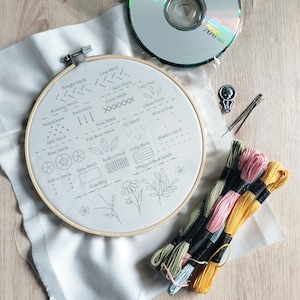 May include: A white embroidery hoop with a variety of embroidery stitches and floral designs drawn on the fabric. The hoop is surrounded by several spools of embroidery floss in various colors.