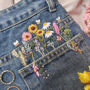 Pu&ograve; includere: Un paio di jeans blu denim con una tasca ricamata con una variet&agrave; di fiori colorati, tra cui girasoli, margherite e fiori rosa. Il ricamo &egrave; dettagliato e intricato, e i fiori sono disposti in un motivo gradevole.