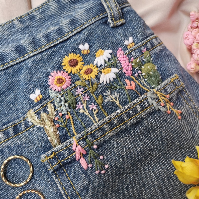 Pu&ograve; includere: Un paio di jeans blu denim con una tasca ricamata con una variet&agrave; di fiori colorati, tra cui girasoli, margherite e fiori rosa. Il ricamo &egrave; dettagliato e intricato, e i fiori sono disposti in un motivo gradevole.