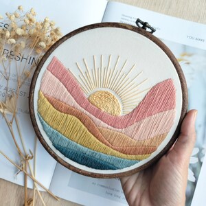 May include: Embroidered hoop art featuring a sunset over a landscape of colorful hills. The sun is yellow with a gold outline and rays, and the hills are shades of pink, orange, yellow, and blue.