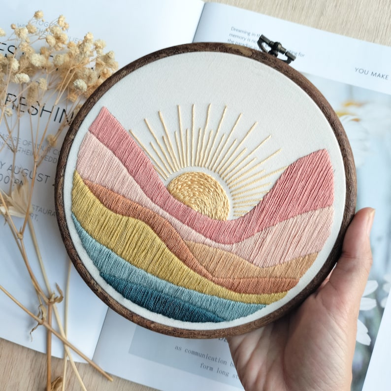 May include: Embroidered hoop art featuring a sunset over a landscape of colorful hills. The sun is yellow with a gold outline and rays, and the hills are shades of pink, orange, yellow, and blue.