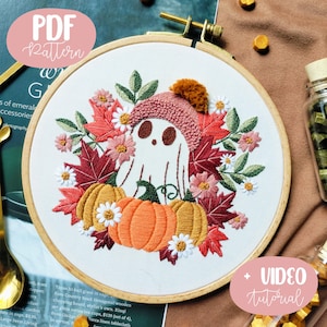 May include: Embroidered hoop art featuring a white ghost wearing a pink hat with a brown pom-pom, sitting on top of two orange pumpkins surrounded by fall leaves and flowers. The text "VIDEO tutorial" is in the lower right corner of the image.