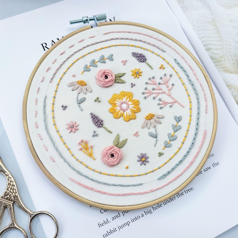 Pu&ograve; includere: Un telaio da ricamo circolare in legno contenente tessuto bianco con disegni floreali cuciti a mano. Il ricamo presenta fiori gialli, rosa e viola, foglie verdi e steli, circondati da bordi circolari concentrici cuciti.