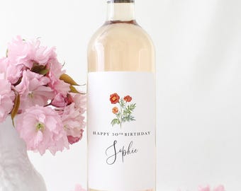 May include: A bottle of rosé wine with a silver cap and a white label featuring a floral design and the text "Happy 30th Birthday Sophie." A vase of pink flowers sits to the left, with scattered pink flowers.