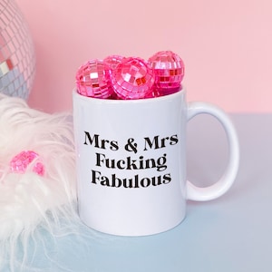 May include: White ceramic mug with a black text design that reads "Mrs & Mrs Fucking Fabulous". The mug is filled with pink disco ball ornaments.