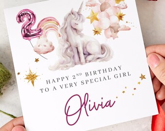 May include: A birthday card featuring a watercolor illustration of a unicorn, balloons, and a rainbow. The card reads "Happy 2nd Birthday to a Very Special Girl" with the name "Olivia" in a cursive font. The card is held by hands.
