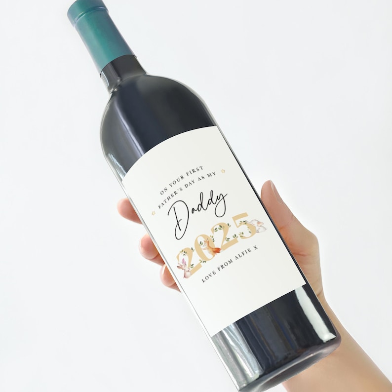 May include: A dark wine bottle with a white label that reads "ON YOUR FIRST FATHER'S DAY AS MY Daddy 2025 LOVE FROM ALFIE X". The bottle has a green top and is held by a person.