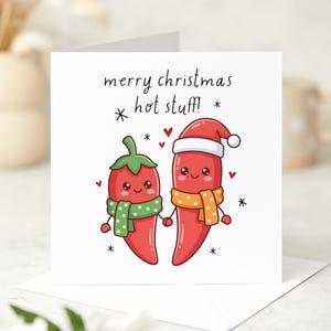 May include: A square white greeting card featuring a cartoon illustration of two red chilli peppers holding hands. The left pepper wears a green polka-dot scarf, and the right wears an orange scarf and a Santa hat. Text reads: merry christmas hot stuff!