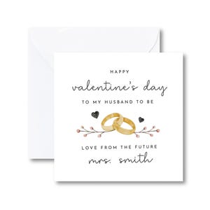 K&ouml;nnte beinhalten: Eine wei&szlig;e Karte mit dem Text "Happy valentine's day to my husband to be" und "Love from the future mrs. smith". Die Karte zeigt eine Aquarell-Illustration von zwei goldenen Eheringen und einem Blumenzweig mit rosa und schwarzen Akzenten.
