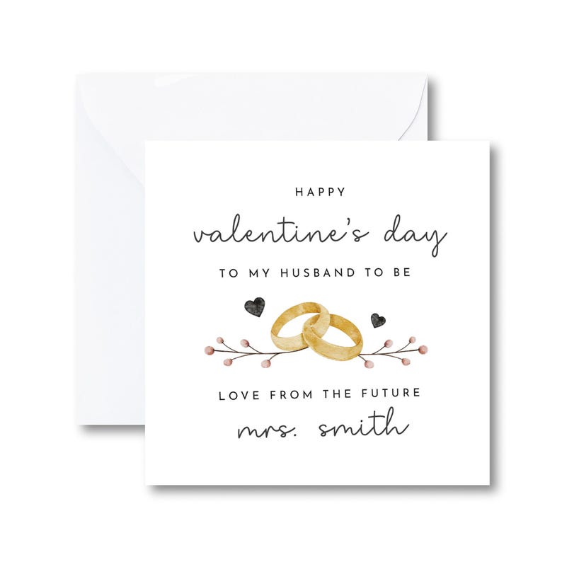 K&ouml;nnte beinhalten: Eine wei&szlig;e Karte mit dem Text "Happy valentine's day to my husband to be" und "Love from the future mrs. smith". Die Karte zeigt eine Aquarell-Illustration von zwei goldenen Eheringen und einem Blumenzweig mit rosa und schwarzen Akzenten.