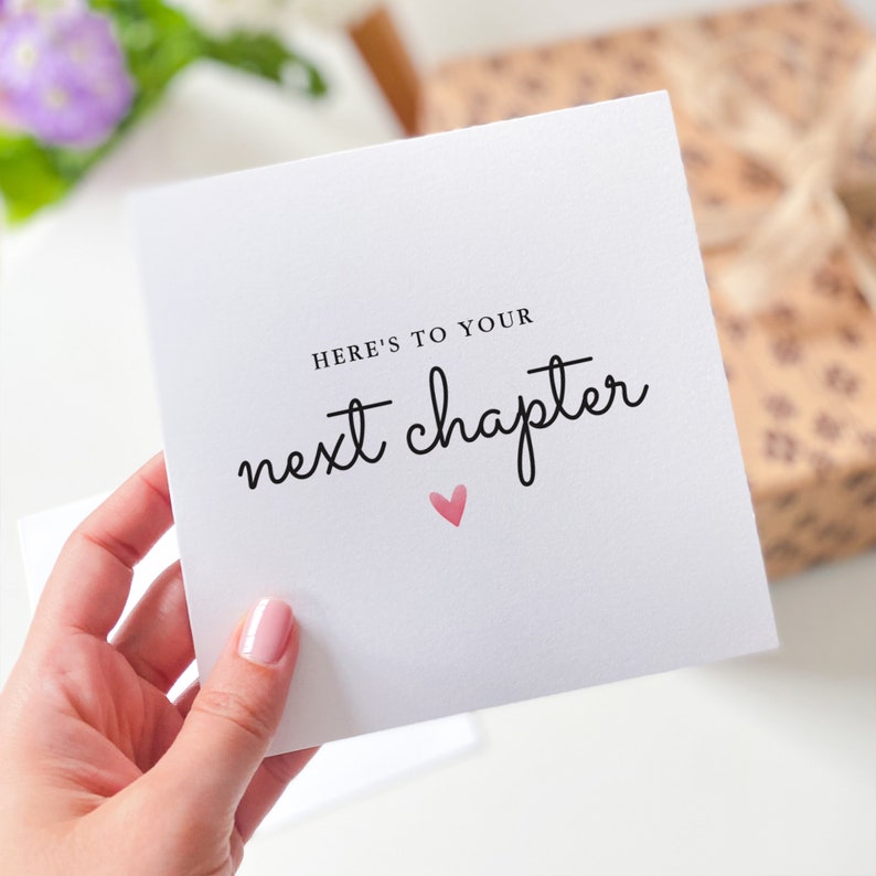 May include: A white card with black text that reads "Here's to your next chapter" with a small pink heart below the text.