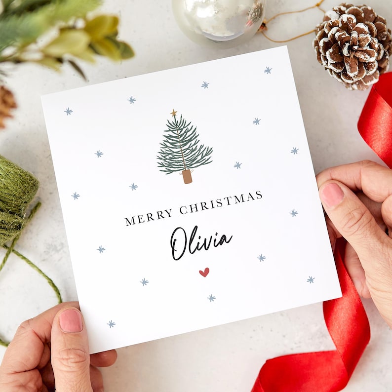 May include: A white Christmas card with a blue and green Christmas tree illustration, a gold star, and the text "Merry Christmas Olivia" with a red heart.