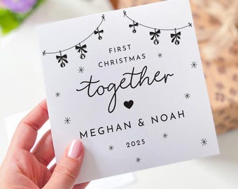May include: A white card with black text and illustrations, held by a hand. The card reads "First Christmas together" with the names "Meghan & Noah" and the year "2025". The design includes bow and star illustrations.