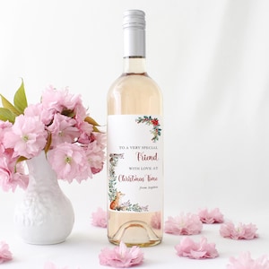 Puede incluir: Una botella de vino blanco con tapón plateado y una etiqueta que dice "Para un amigo muy especial con amor en Navidad". La etiqueta presenta ilustraciones florales y de zorro. Un jarrón blanco contiene flores rosas.