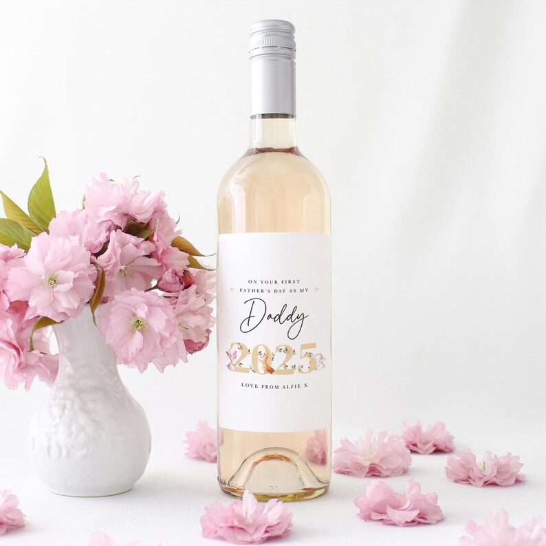 May include: A bottle of ros&eacute; wine with a white label that reads "On your first Father's Day as my Daddy 2025 Love from Alfie". The bottle is next to a white vase with pink flowers and scattered pink flowers.