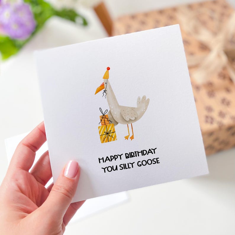 Puede incluir: Una tarjeta blanca con un dibujo animado de un ganso con un gorro de fiesta y sosteniendo una pila de regalos envueltos. El texto de la tarjeta dice "Feliz cumplea&ntilde;os, tonto ganso".