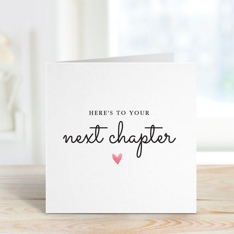 May include: White card with black text that reads "Here's to your next chapter" with a pink heart below the text.