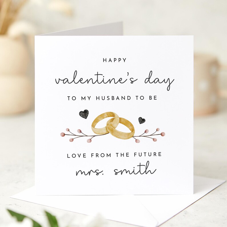 K&ouml;nnte beinhalten: Eine wei&szlig;e Karte mit dem Text "Happy Valentine's Day to my husband to be" und "Love from the future Mrs. Smith". Die Karte zeigt eine Aquarell-Illustration von zwei goldenen Ringen und einem Blumenzweig.
