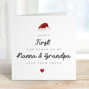 May include: A white Christmas card with a Santa hat illustration. The card reads "Happy First Christmas as my Nanna & Grandpa" in black script, with "Love from Freya" below a red heart.