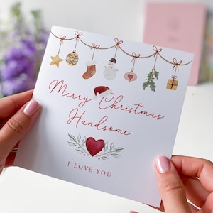 May include: A white Christmas card with a red heart and the text "Merry Christmas Handsome I Love You". The card is decorated with a string of Christmas ornaments, including a star, a stocking, a snowman, and a present.