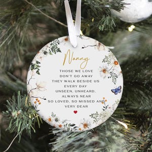 May include: A white, round Christmas ornament with a floral wreath design. The ornament has the word "Nanny" in gold script and a poem. A small blue butterfly and a red heart are also featured.