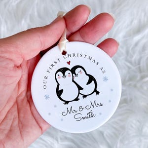 Könnte beinhalten: Ein weißer Keramik-Weihnachtsschmuck mit cremefarbenem Band. Der Schmuck zeigt zwei Cartoon-Pinguine, Schneeflocken und den Text "Our First Christmas As Mr & Mrs Smith."