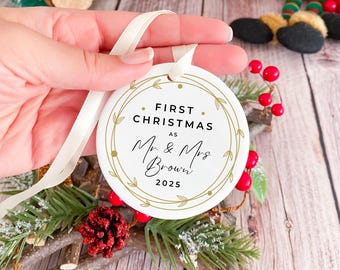 May include: A white, round Christmas ornament with gold lettering and a gold leaf design. The text reads "FIRST CHRISTMAS as Mr & Mrs Brown 2025". A cream-colored ribbon is attached for hanging. The ornament is a festive holiday decoration.