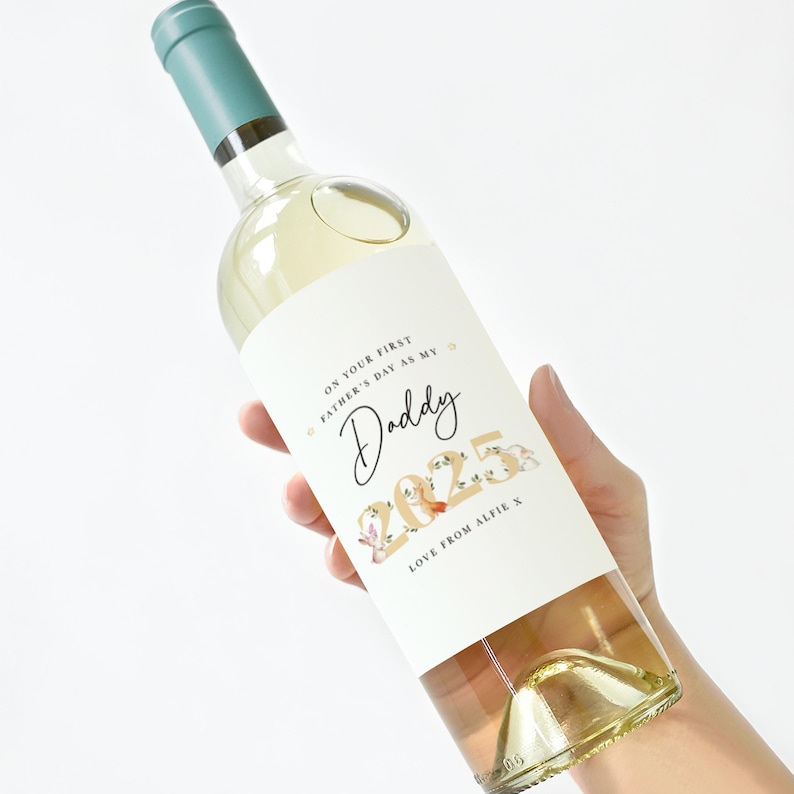 May include: A clear wine bottle with a teal cap and a white label. The label reads "ON YOUR FIRST FATHER'S DAY AS MY Daddy 2025 LOVE FROM ALFIE X" in black and gold script with floral accents. The bottle is filled with a light-colored liquid.