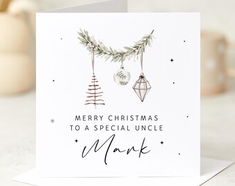 May include: A white Christmas card with a watercolor design. The card features a green garland with hanging ornaments, including a tree, a ball, and a geometric shape. The text reads "MERRY CHRISTMAS TO A SPECIAL UNCLE" and "Mark".