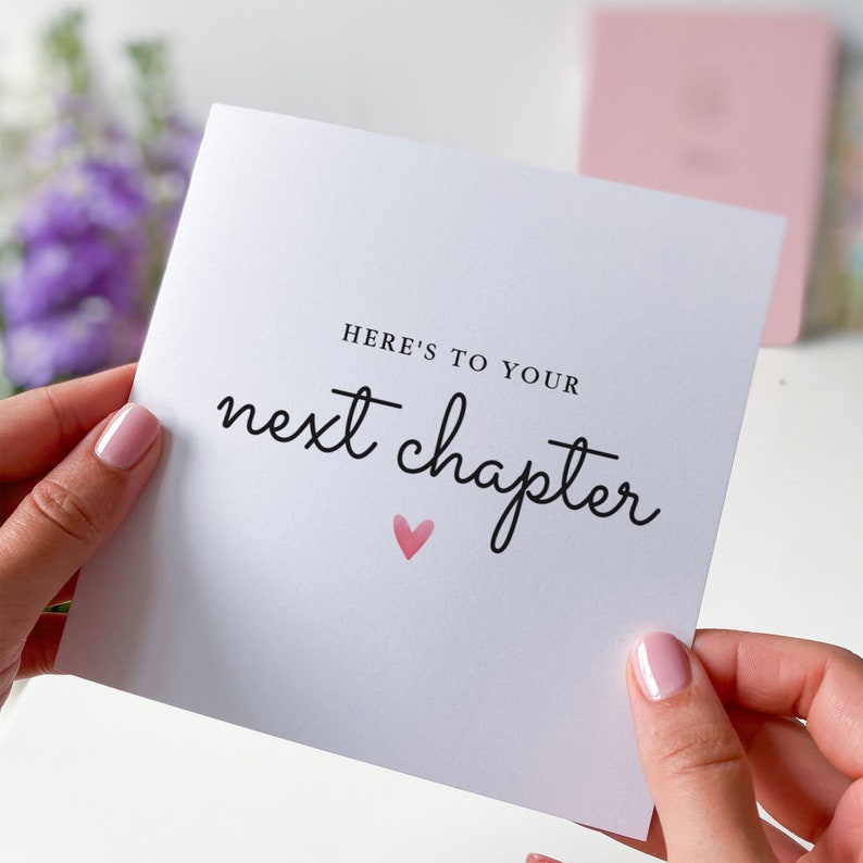 May include: White card with black text that reads "Here's to your next chapter" with a pink heart.