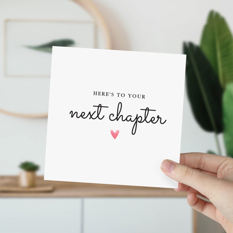 May include: White card with black text that reads "Here's to your next chapter" with a pink heart below the text.