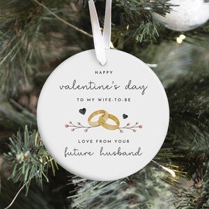 Può includere: Un ornamento rotondo bianco di San Valentino con un nastro bianco. L'ornamento presenta il testo "Happy Valentine's Day To My Wife-To-Be" e "Love From Your Future Husband". Sono inoltre visualizzati anelli nuziali d'oro e cuori.