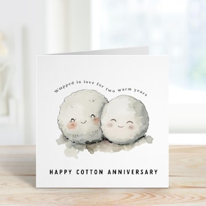Könnte beinhalten: Eine weiße Grußkarte mit einer Aquarellillustration von zwei lächelnden Wattebällchen-Charakteren. Die Karte trägt den Text "Wrapped in love for two warm years" und "Happy Cotton Anniversary" in schwarzer Schrift.