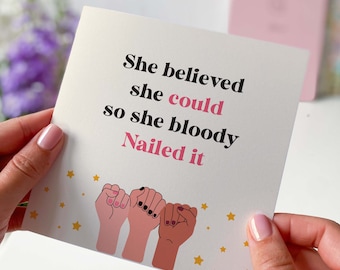 May include: A white card with the text "She believed she could so she bloody nailed it" in black and pink lettering. Three stylized fists in pink, brown, and peach are shown at the bottom of the card.