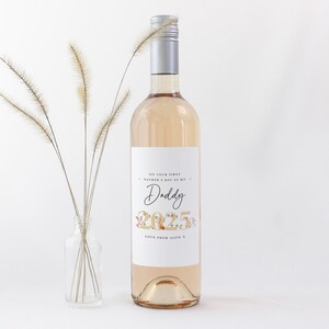 May include: A clear wine bottle with a silver cap and a white label. The label reads "ON YOUR FIRST FATHER'S DAY AS MY Daddy 2025 LOVE FROM ALFIE X". A small glass vase with dried flowers is to the left.