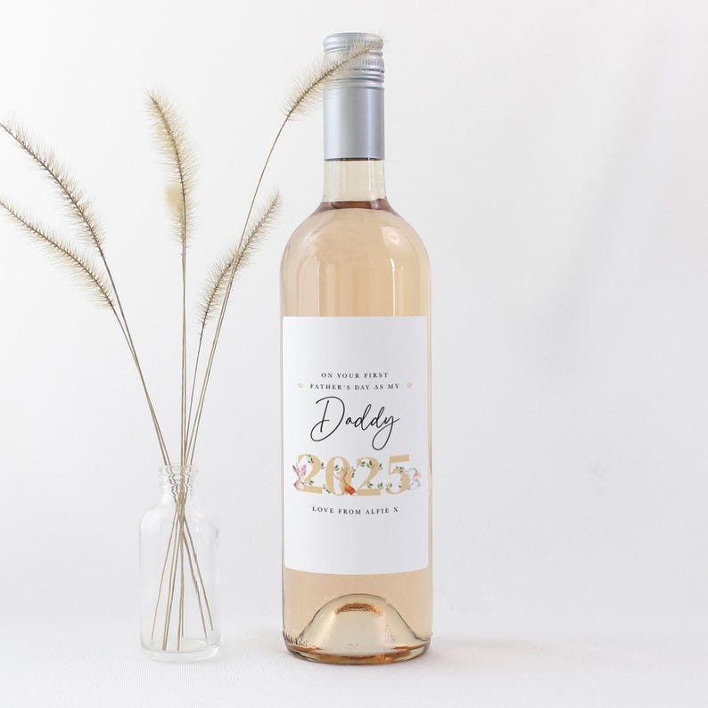 May include: A clear wine bottle with a silver cap and a white label. The label reads "ON YOUR FIRST FATHER'S DAY AS MY Daddy 2025 LOVE FROM ALFIE X". A small glass vase with dried flowers is to the left.