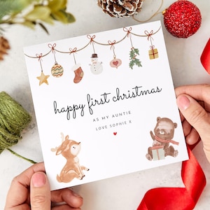 May include: A white card with a red heart and the text "happy first christmas as my auntie love sophie x". The card is decorated with a string of Christmas ornaments, including a snowman, a stocking, a star, a bauble, a heart, and a sprig of holly. There are also illustrations of a deer and a bear.