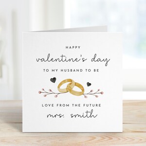 K&ouml;nnte beinhalten: Eine wei&szlig;e Karte mit schwarzem Text, der "Happy valentine's day to my husband to be Love from the future mrs. smith" lautet. Die Karte zeigt eine Aquarell-Illustration von zwei ineinander verschlungenen goldenen Ringen mit einem Zweig mit rosa Beeren und schwarzen Herzen.
