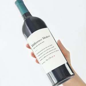 Op de afbeelding: Een donkere wijnfles met een wit etiket met de tekst "Difference Maker". De fles heeft een groene bovenkant en wordt schuin gehouden. Het etiket definieert "Difference Maker" als een persoon die een grote impact heeft.